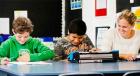 A teacher aide works alongside a student who is using a braillewriter machine with another student who is writing notes
